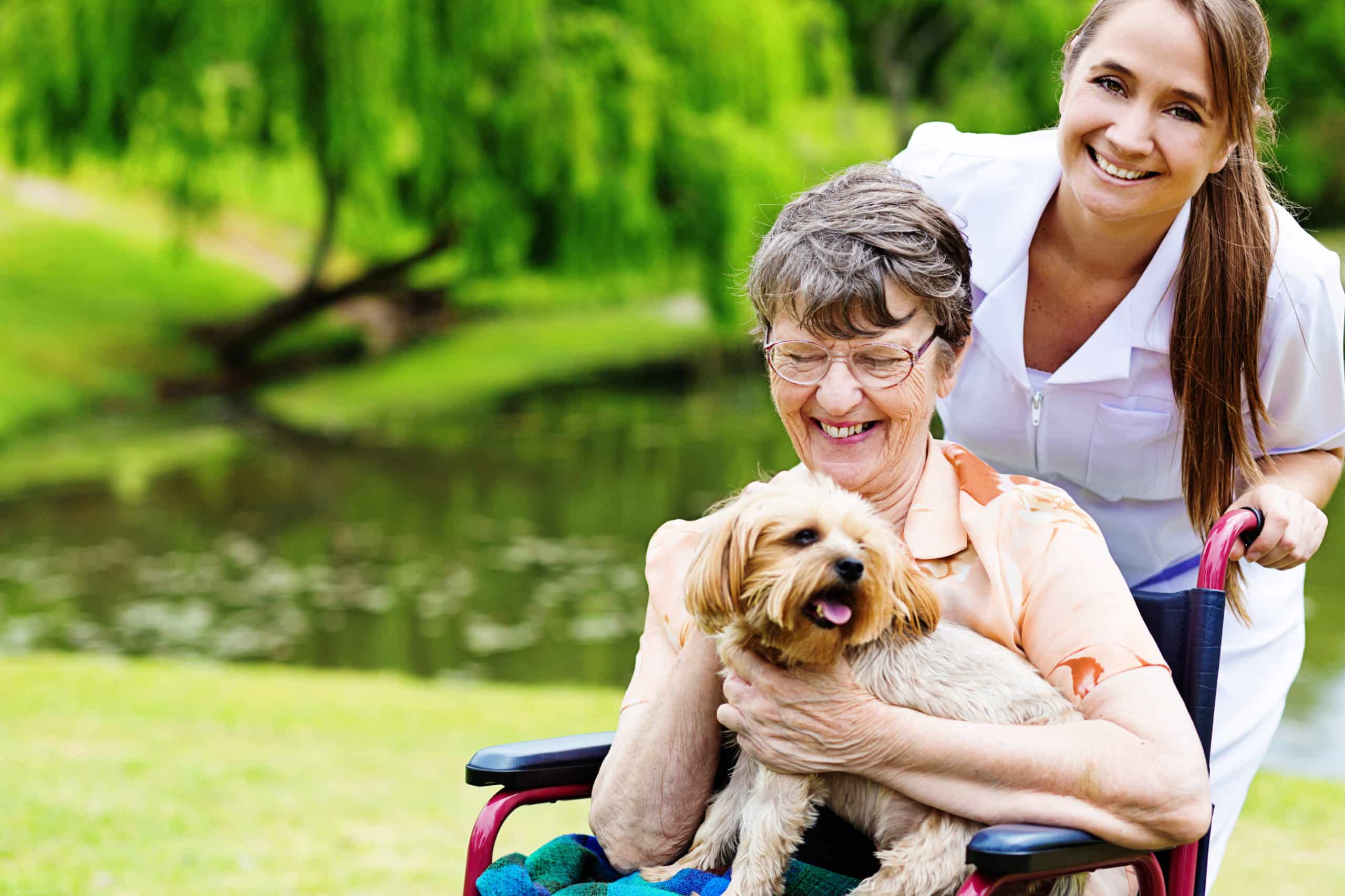 Happy old wheelchair-bound woman with pet dog and smiling caregiver
