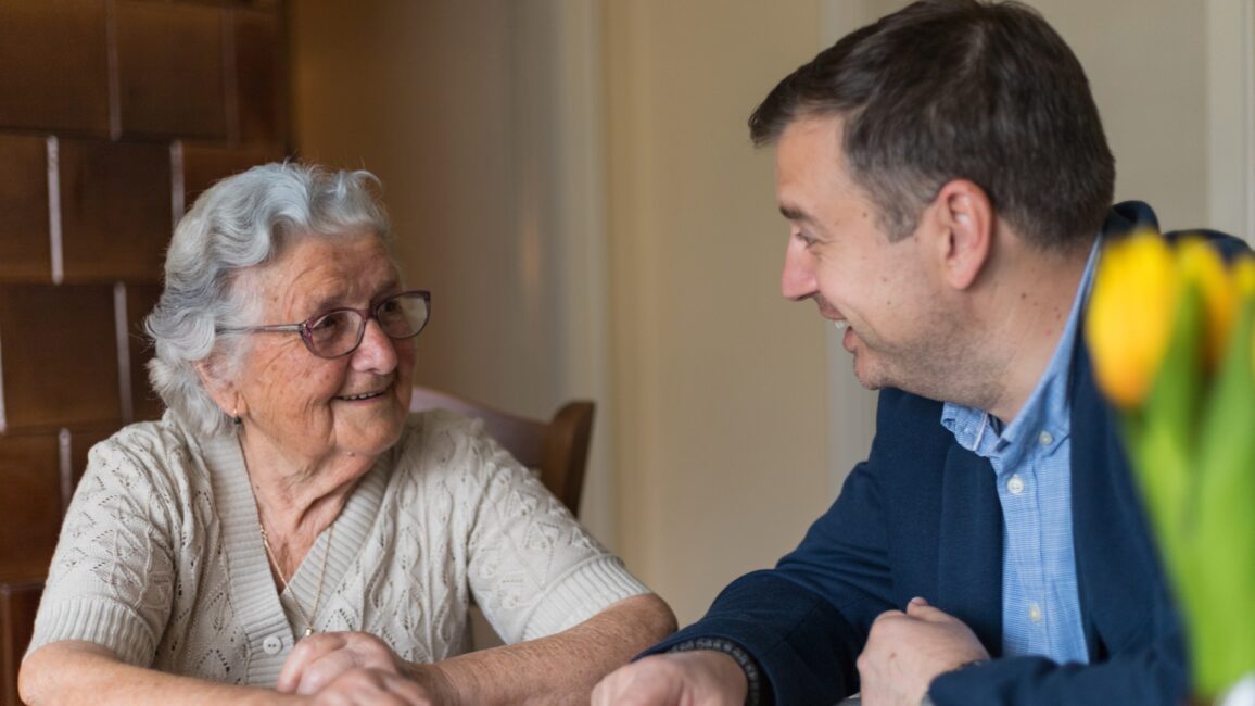 Senior woman with her adult grandson are looking old photos and remember the old memories.Man enjoys the company of his grandmother and spending wonderful time with her. Old woman with gray hair, glasses and a middle-aged man in a jacket are sitting at the table and looking at the photos in the old family house.