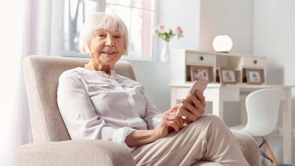 Advanced user. Cheerful senior woman sitting in an armchair and smiling at the camera while surfing the Internet on her phone
